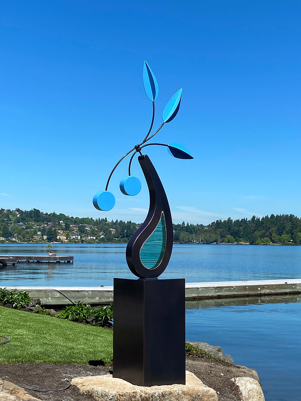 bronze kidney shaped sculpture with blue kinetic arms on a lake