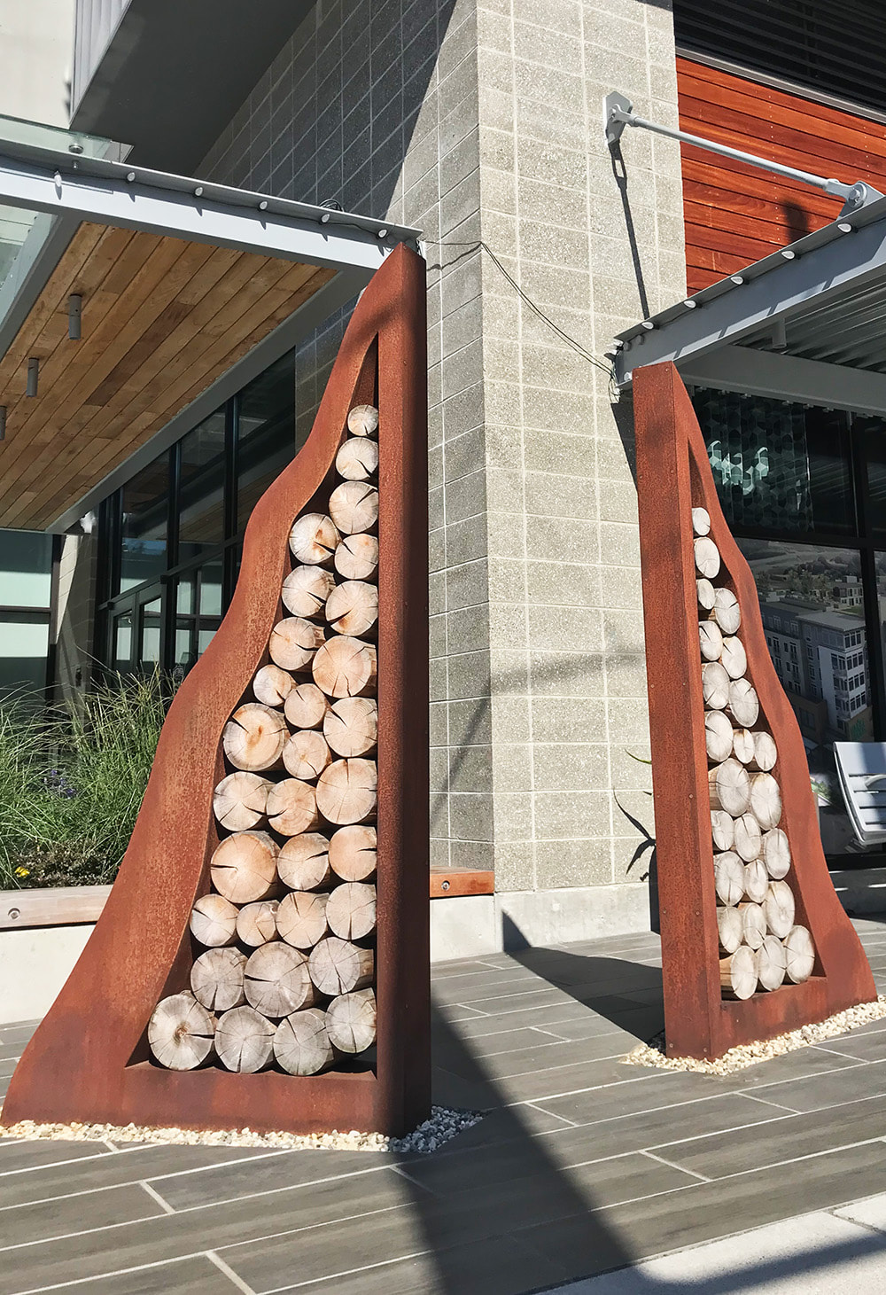 corten sculpture with cedar logs inlayed in front of a multi use building outside